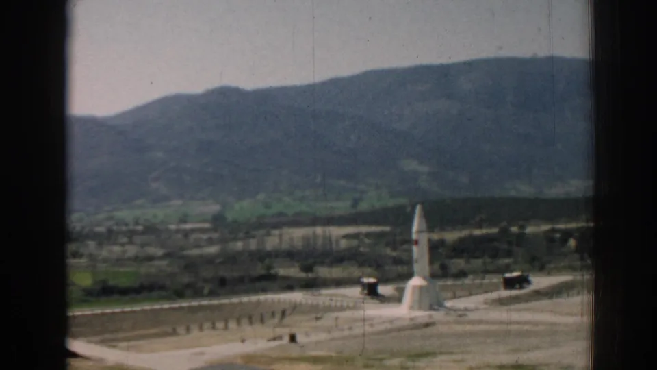 1962 Izmir-region home-movie frame showing a white rocket-like monument, pale sky, roads, hills, and distant mountains through a dark window frame.