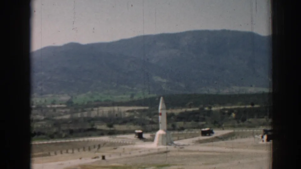 1962 Izmir-region view through a dark window frame toward a white monument set near roads, fields, green hills, and a mountain ridge.