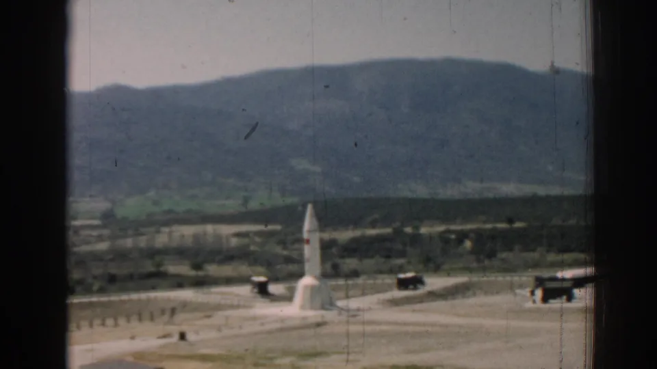 1962 Izmir-region home-movie view of a white monument, low roadside structures, sandy ground, hills, and mountains seen through a window frame.