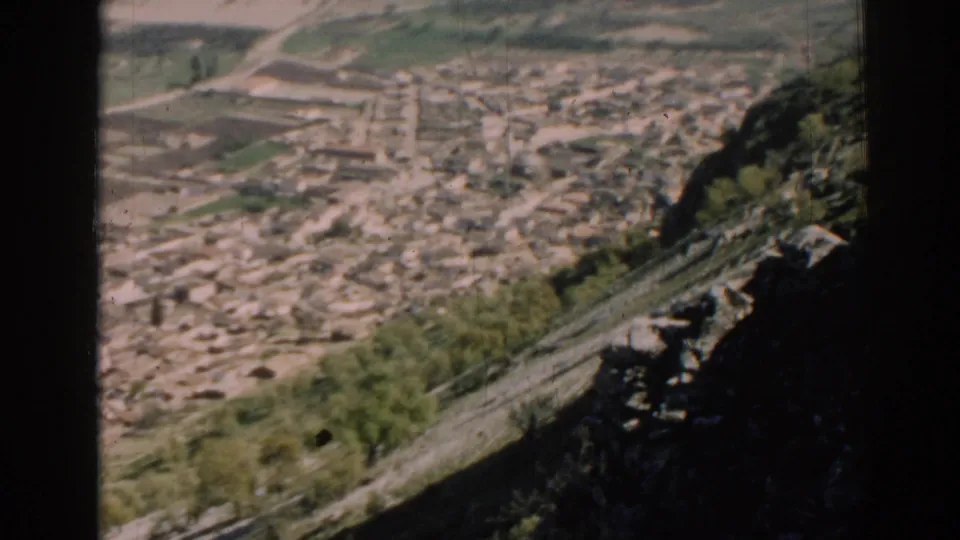 1962 Izmir-region frame showing an elevated view over a compact valley town, winding roads, pale terrain, and a rocky hillside edge.