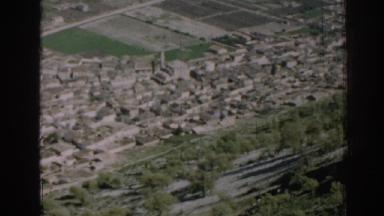 Elevated view of low-rise buildings, fields, trees, and a road on a hillside.
