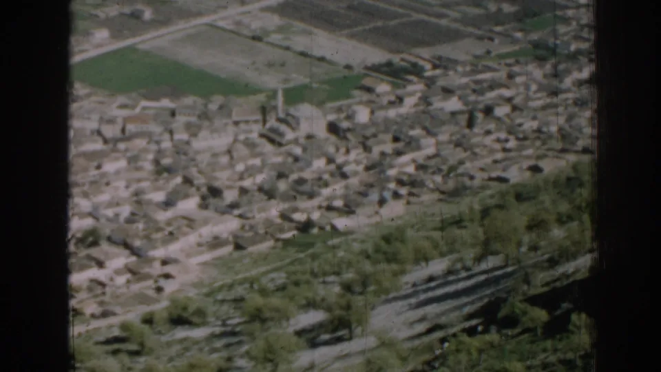 1962 Izmir home-movie frame showing dense low-rise buildings beyond green fields, with trees and a hillside road in the foreground.