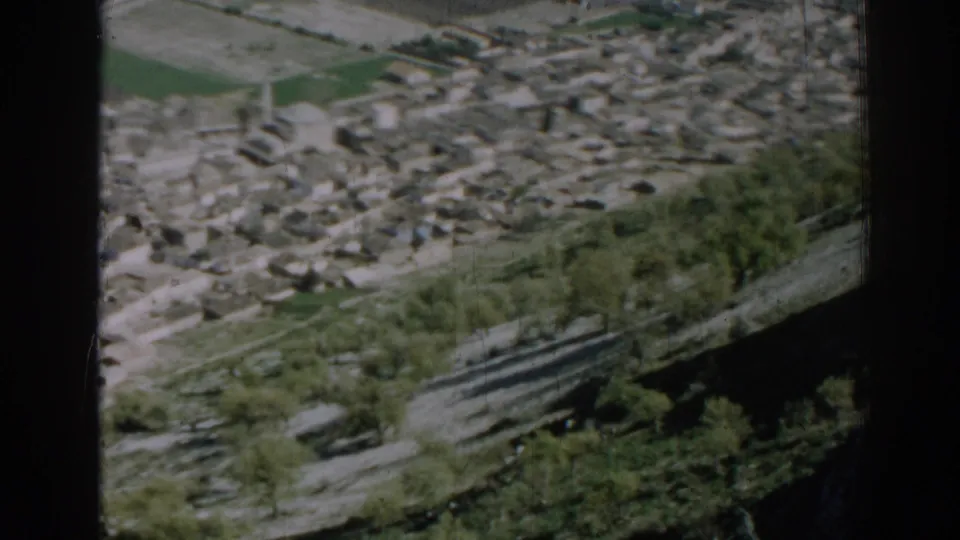 1962 Izmir frame with clustered pale buildings, cultivated green plots, hillside trees, and a curving concrete road below.