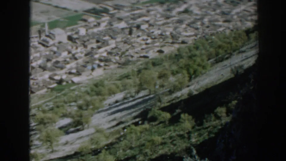 Soft 1962 Izmir frame showing a dense settlement beyond trees and road surfaces, with dark hillside shadows along the lower edge.