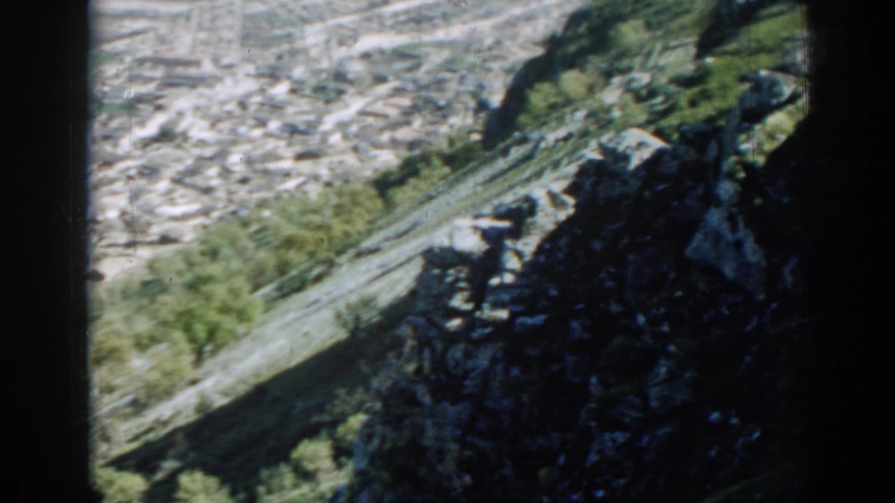 1962 8mm view of an Izmir hillside settlement, with clustered rooftops beyond a rocky tree-lined slope.