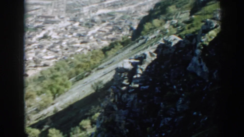 1962 8mm view of an Izmir hillside settlement, with clustered rooftops beyond a rocky tree-lined slope.