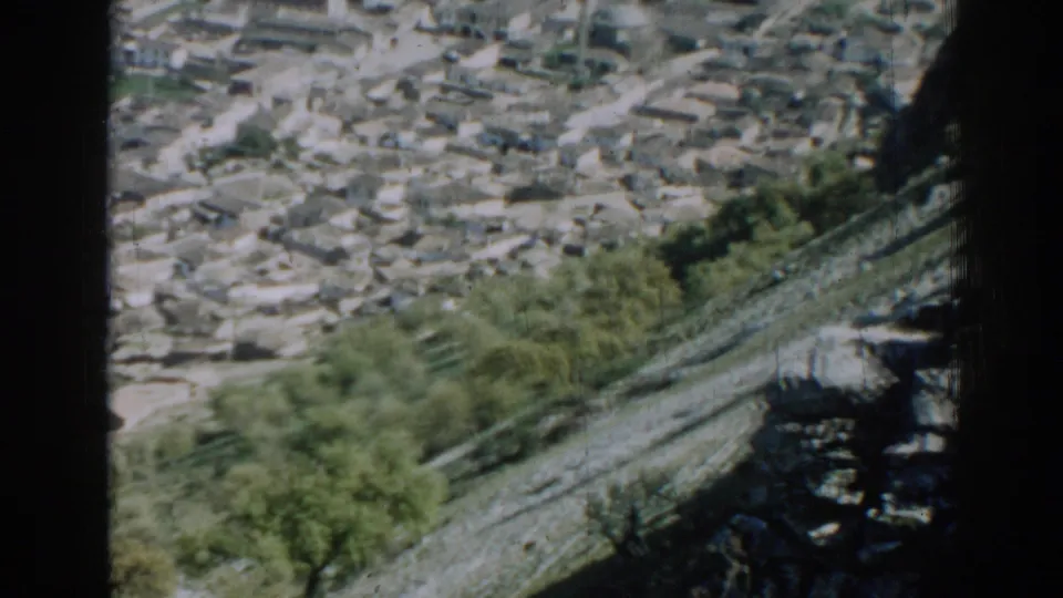 A grainy 1962 home-movie frame shows Izmir rooftops, trees, and paths from an elevated hillside viewpoint.