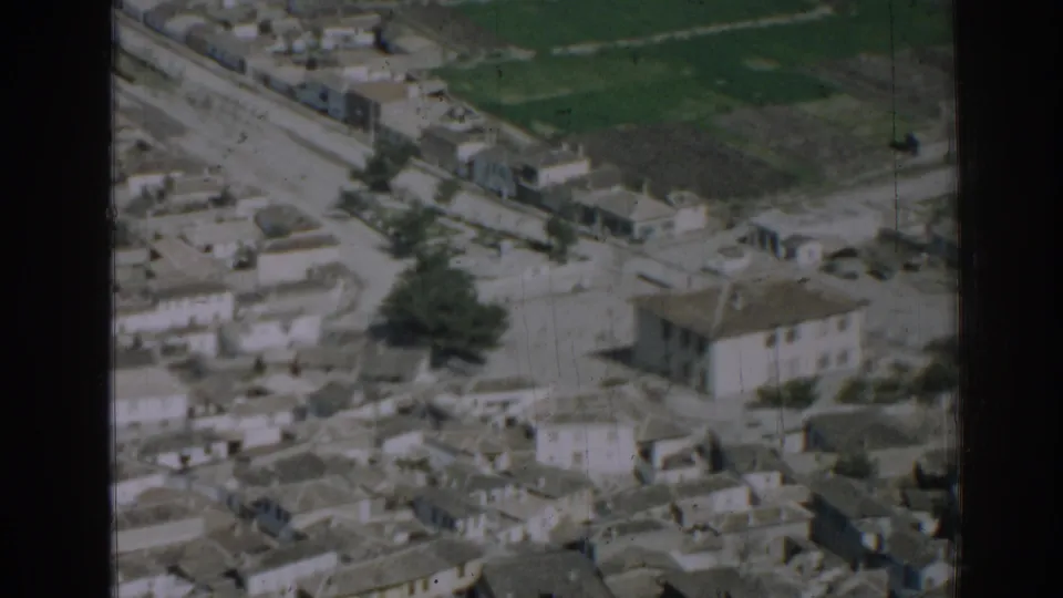 1962 Izmir aerial view passing over a larger two-story building among dense rooftops and nearby fields.