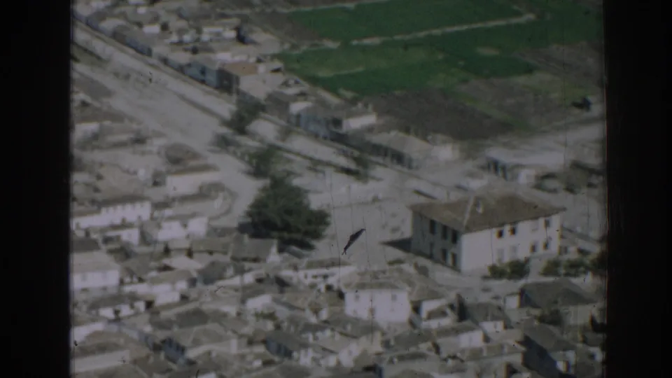 1962 Izmir aerial frame showing a larger building amid clustered homes, pale streets, green fields, and 8mm wear.
