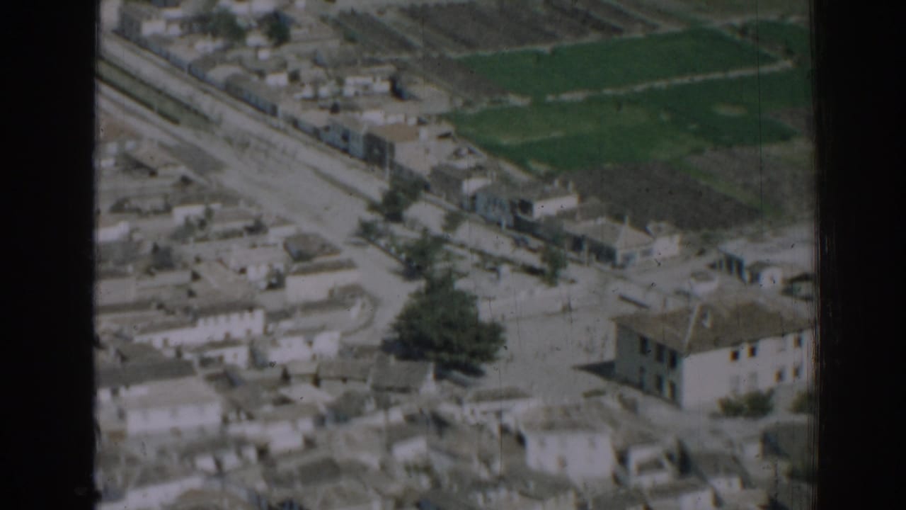 Aerial view of low-rise buildings, a road, railway line, and green fields.