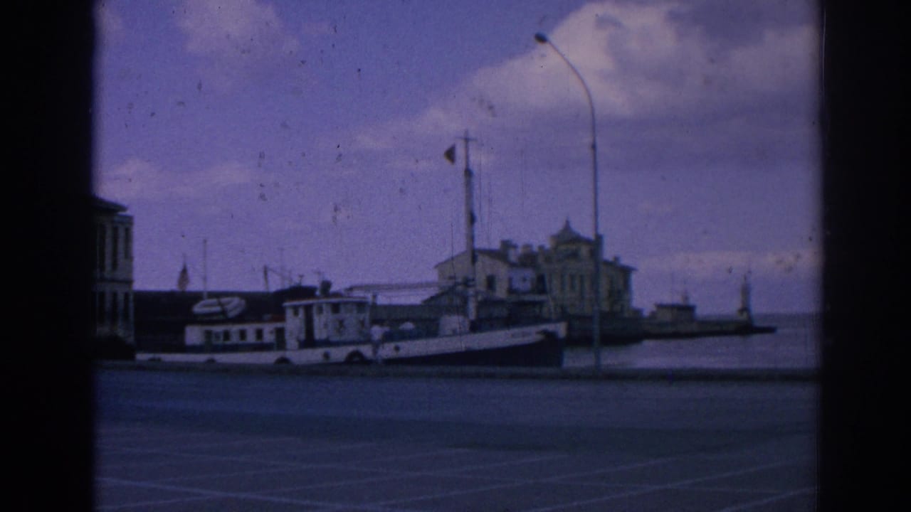 White boat docked beside a quay with a red flag, lamppost, waterfront building, and cloudy sky.