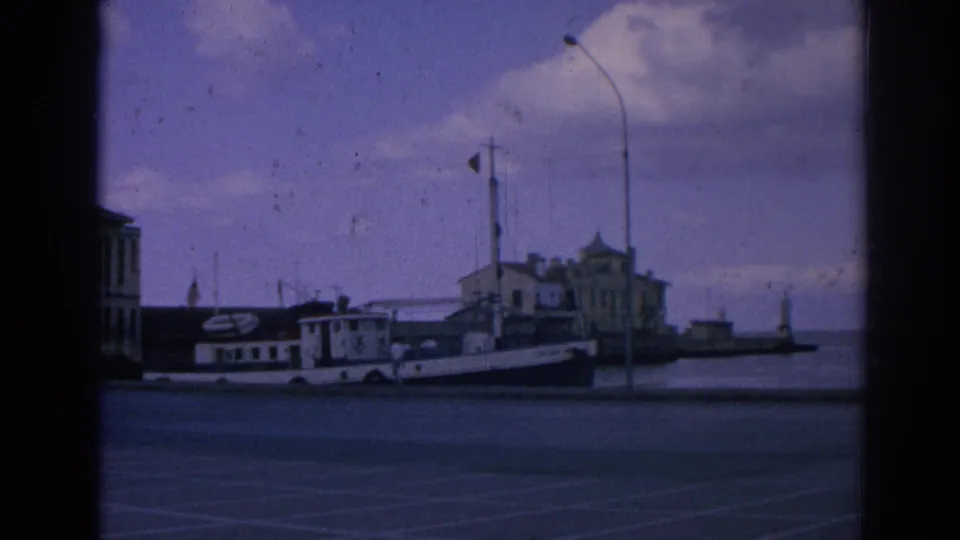 1962 8mm view of a white docked boat at an Izmir waterfront, with a red flag, lamppost, harbor water, and buildings beyond.