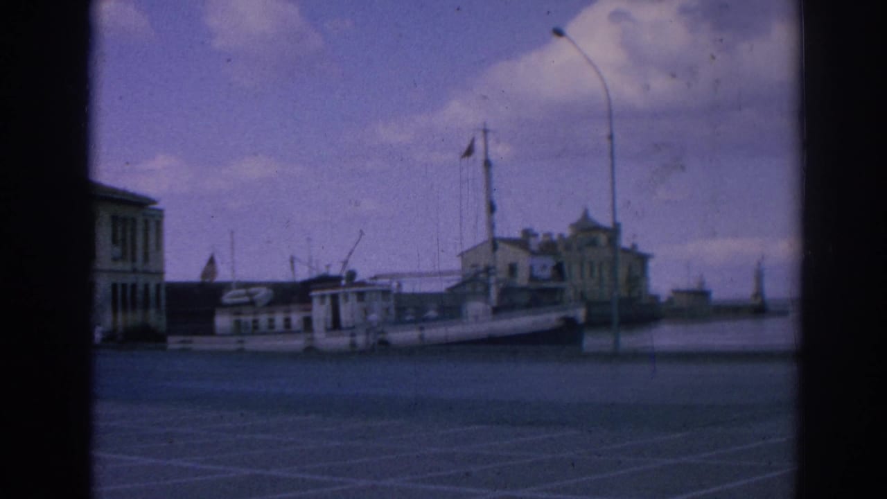Docked white boat near harbor buildings, with a red flag, lamppost, quay, water, and sky.