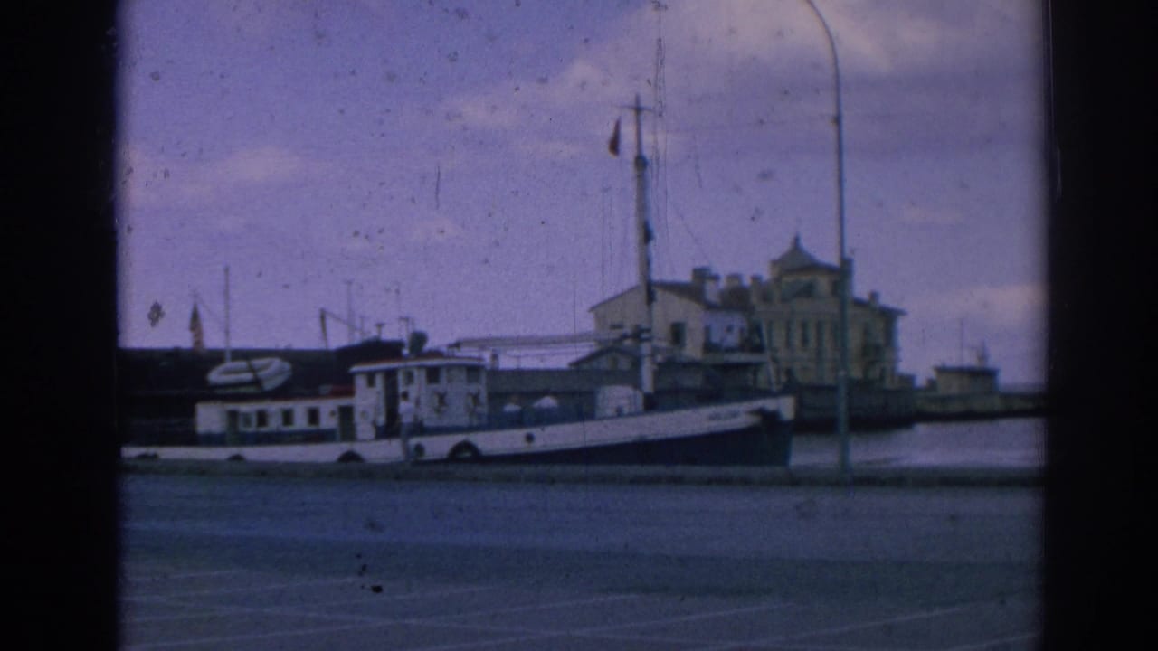 Closer 1962 8mm frame of the Izmir waterfront with a white moored boat, red flag on a mast, lamppost, quay, and harbor buildings.