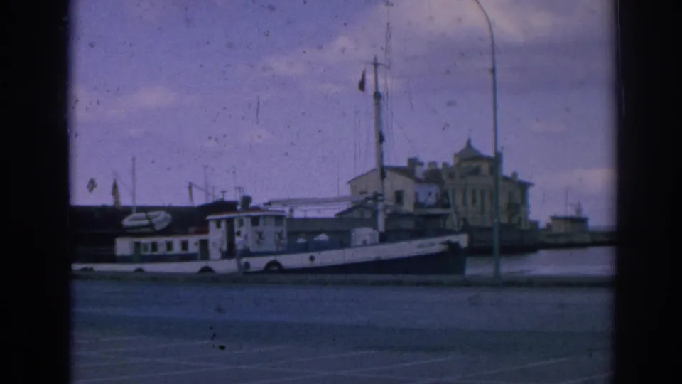 Closer 1962 8mm frame of the Izmir waterfront with a white moored boat, red flag on a mast, lamppost, quay, and harbor buildings.