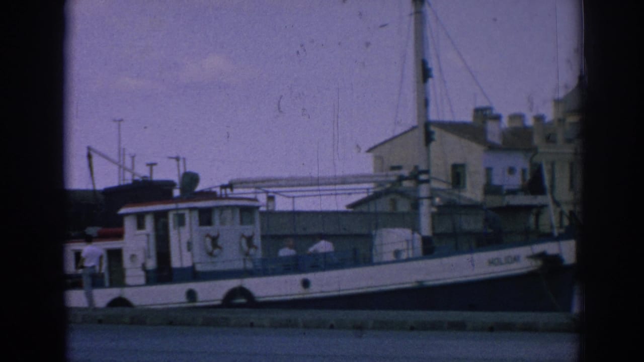 1962 Izmir home-movie frame showing the tugboat HOLIDAY docked near waterfront buildings under a muted sky.
