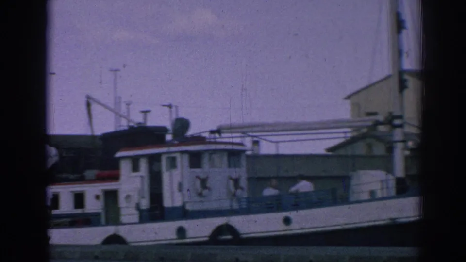Side view of the white and blue tugboat HOLIDAY at a marina in Izmir, Turkey, with cabin details and dock structures visible.