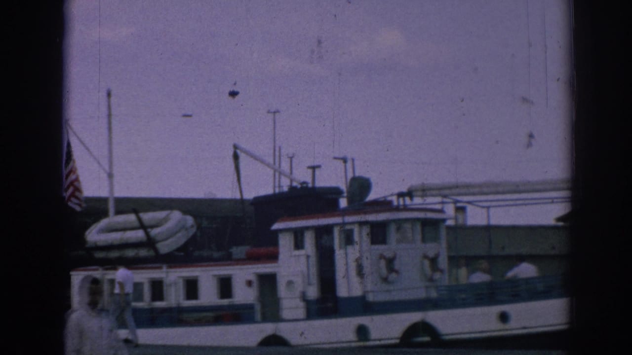 The docked tugboat is framed with an American flag, a large inflatable tube, and mid-century harbor equipment in the 1962 Izmir footage.