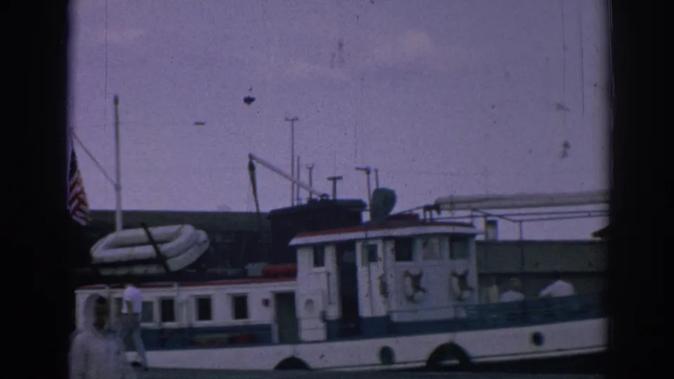 The docked tugboat is framed with an American flag, a large inflatable tube, and mid-century harbor equipment in the 1962 Izmir footage.