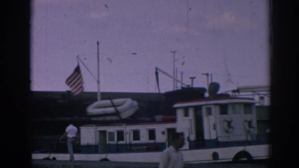 Final frame of the Izmir marina scene shows the docked tugboat, American flag, people on or near the pier, and a man passing in the foreground.