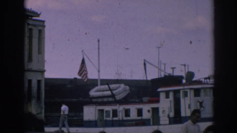 1962 Izmir waterfront view with a docked boat, American flag, building edge, and a man walking along the quay.