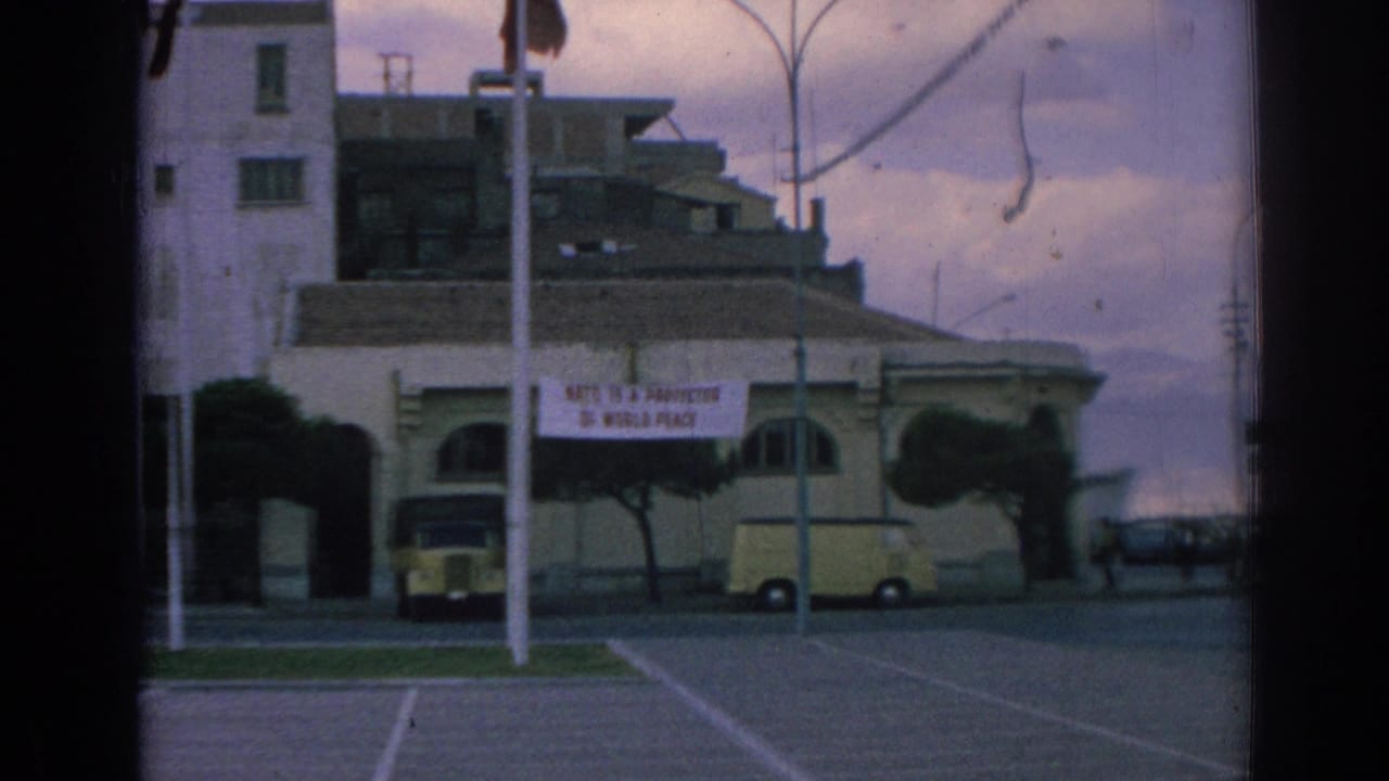Izmir street-side building displaying a NATO peace banner, with a yellow van, parked car, poles, and trimmed trees in view.