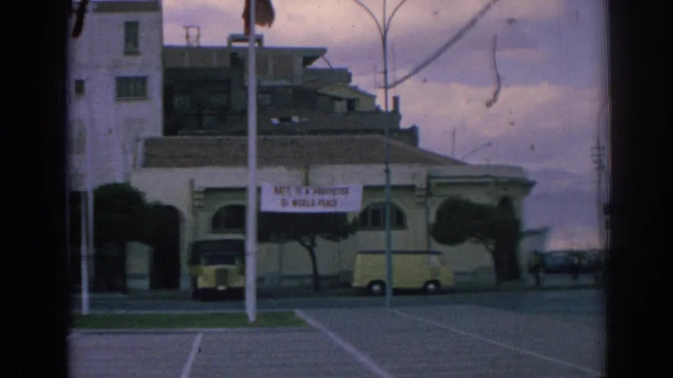 Izmir street-side building displaying a NATO peace banner, with a yellow van, parked car, poles, and trimmed trees in view.