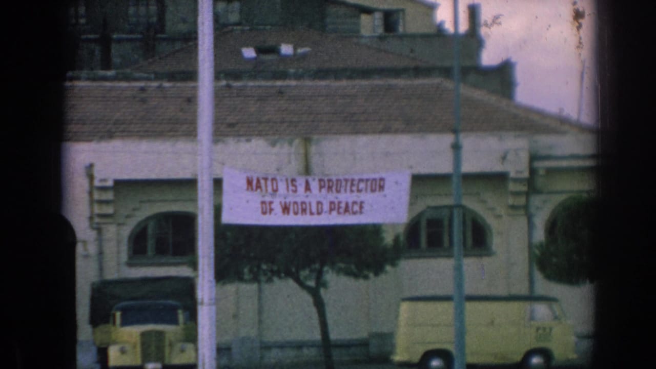 Closer 1962 Izmir view of a banner reading NATO IS A PROTECTOR OF WORLD PEACE on a building above parked vehicles.