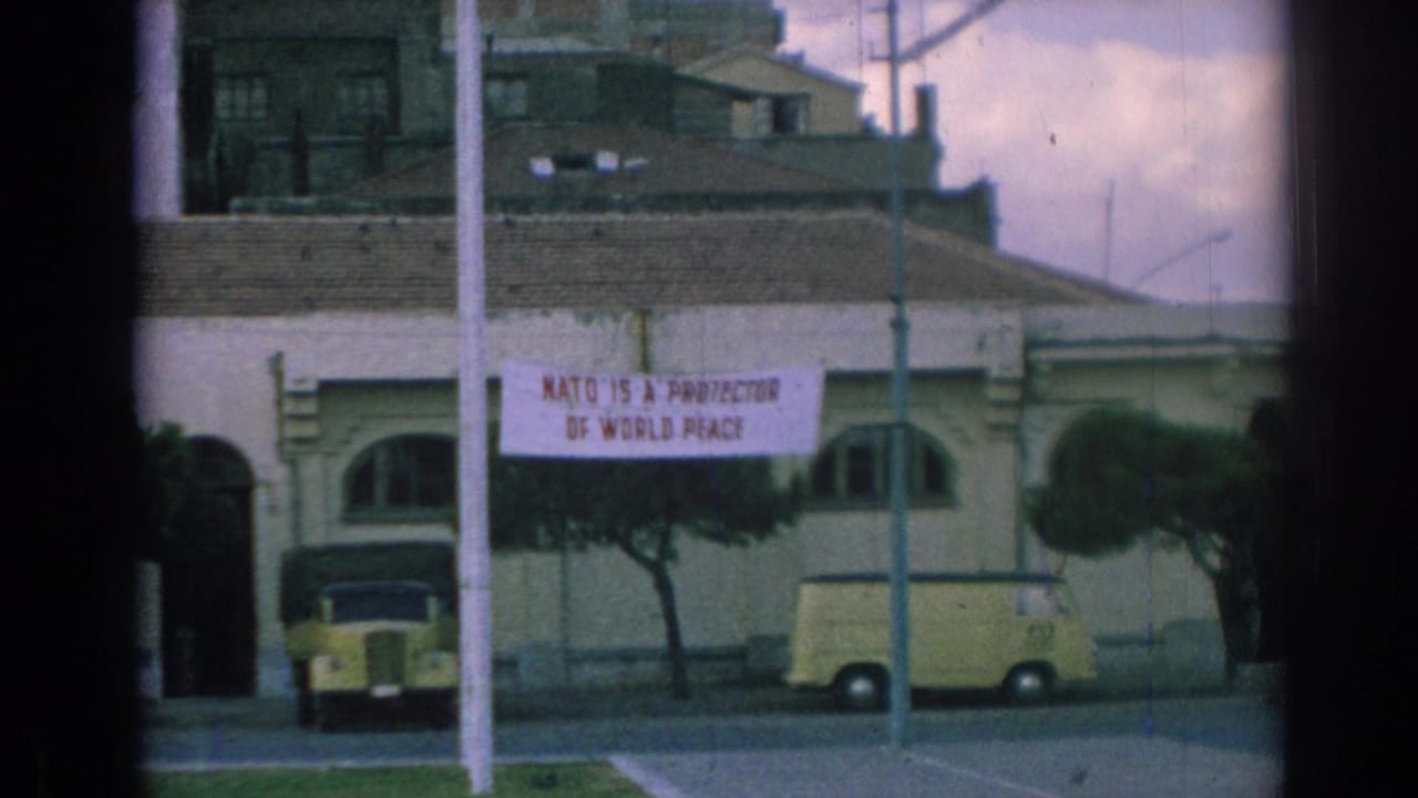 Building with arched windows, NATO peace banner, yellow car, and yellow van on a street.