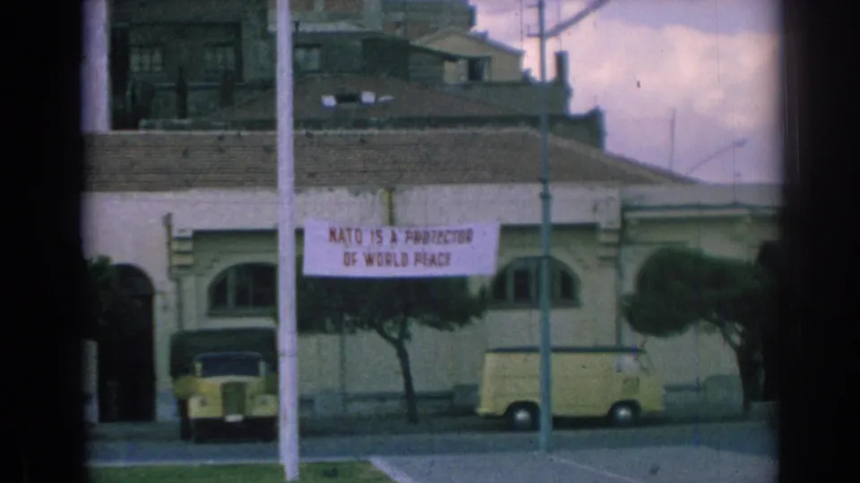 1962 Izmir street view with a pale building, red-lettered NATO peace banner, parked yellow car, yellow van, trees, and street poles.