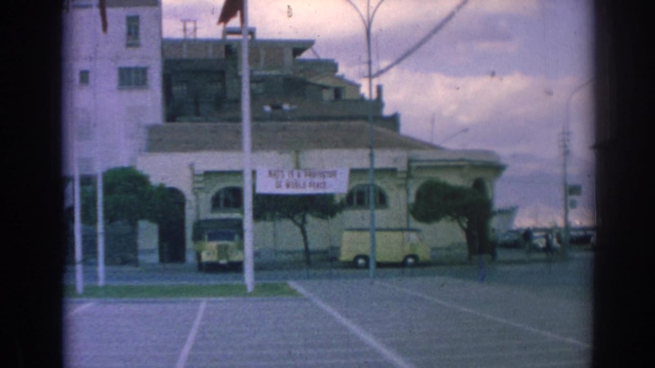 Wider 1962 Izmir view showing the NATO banner on a pale building, yellow van and car parked nearby, streetlights, trees, and an open paved area.