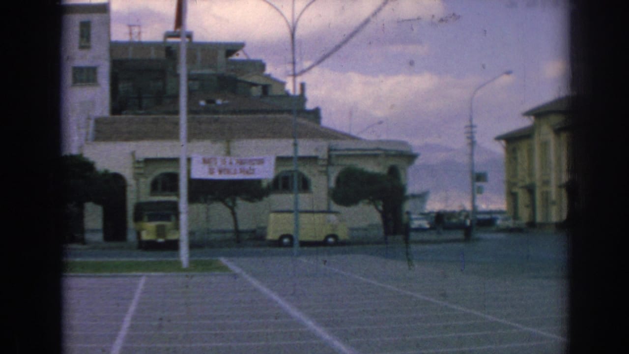 1962 Izmir street scene with a NATO peace banner, pale building facade, parked yellow vehicles, open lot markings, street poles, and cloudy sky.