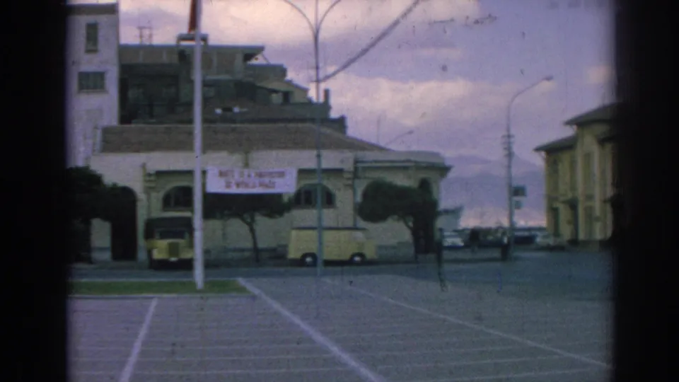 1962 Izmir street scene with a NATO peace banner, pale building facade, parked yellow vehicles, open lot markings, street poles, and cloudy sky.