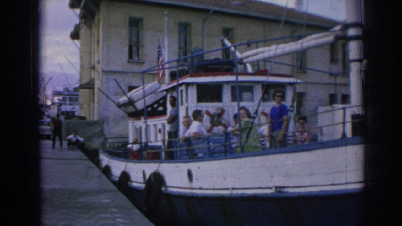1962 Izmir harbor-side view of a white houseboat tied at a pier, with adults visible on deck, flags, railings, water, and waterfront buildings.