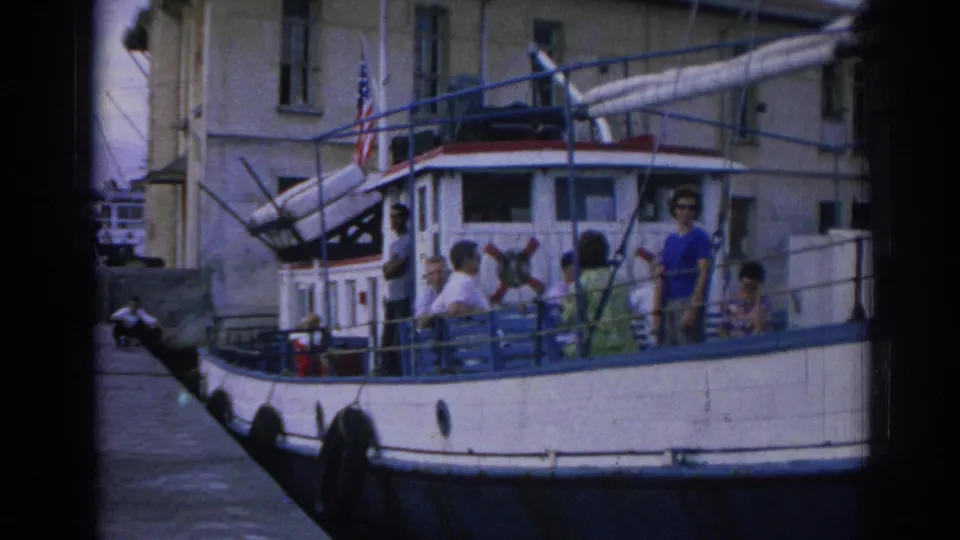 1962 Izmir home-movie frame showing passengers on a white and blue boat, with an American flag and waterfront building behind them.