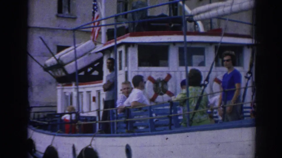 1962 Izmir frame with several passengers on a small boat deck, including a woman in blue sunglasses near a red life preserver.