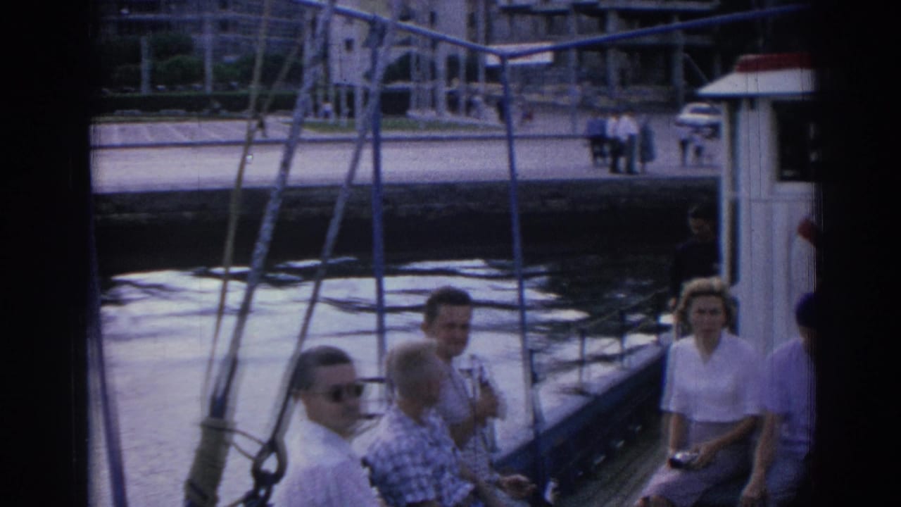 Passengers sit on a tour boat near an urban waterfront with rigging in the foreground.