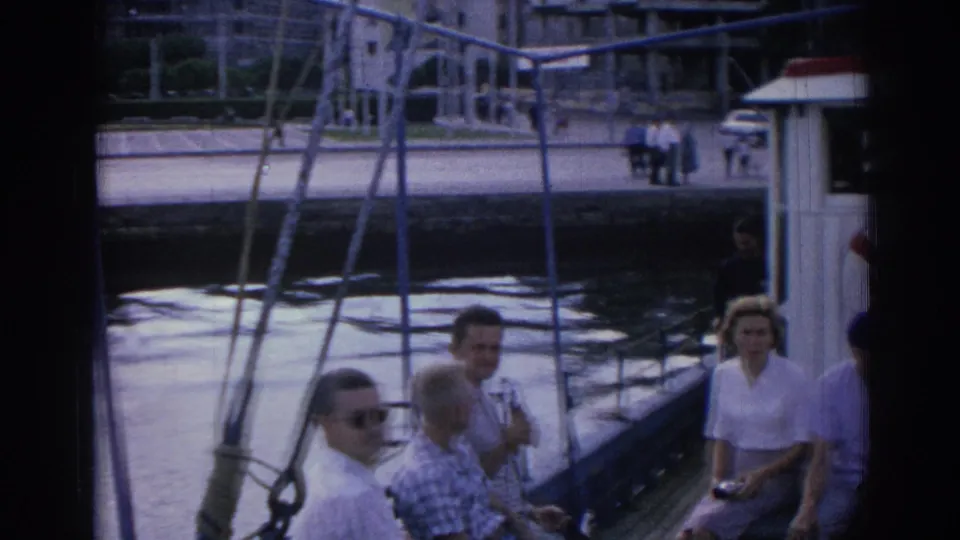1962 Izmir tour boat passengers sit near rigging as the camera looks toward a paved waterfront and modern buildings.
