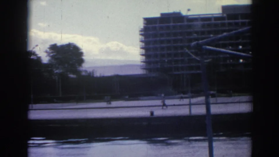 The camera looks from the boat across water toward an Izmir promenade, trees, and a large building under construction.