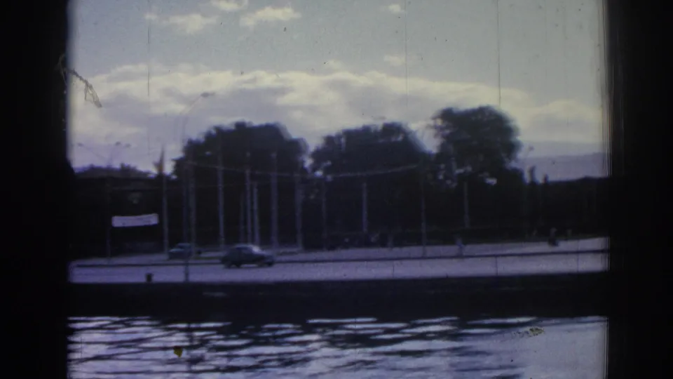 1962 Izmir waterfront street seen across water, with cars passing near trees, flagpoles, and a pale banner.