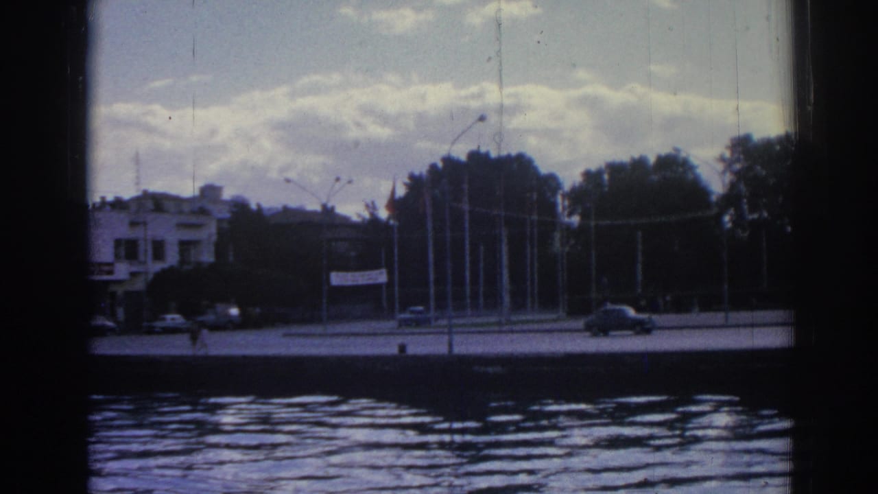 Across-water view of an Izmir street in 1962, with cars, pedestrians, flagpoles, trees, and a low building in the background.