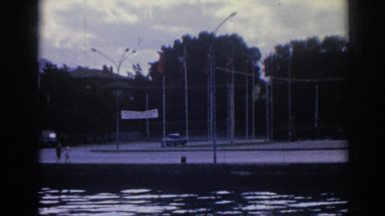 1962 Izmir waterfront scene with a car, distant pedestrians, flagpoles, streetlamps, trees, and a white banner by the road.