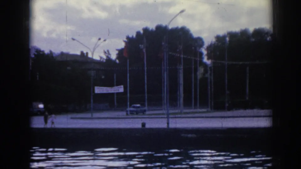 1962 Izmir waterfront scene with a car, distant pedestrians, flagpoles, streetlamps, trees, and a white banner by the road.