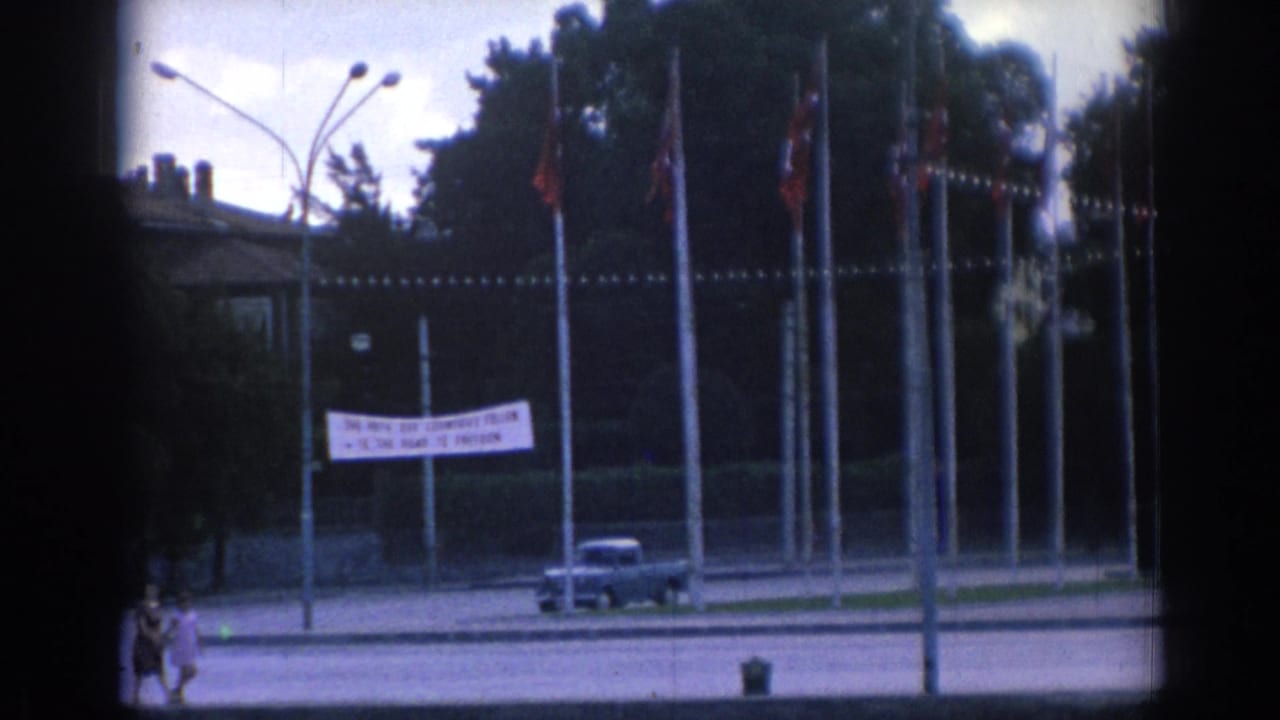 Closer 1962 Izmir view of a waterfront road with flagpoles, a white banner, a car, trees, streetlamps, and distant pedestrians.