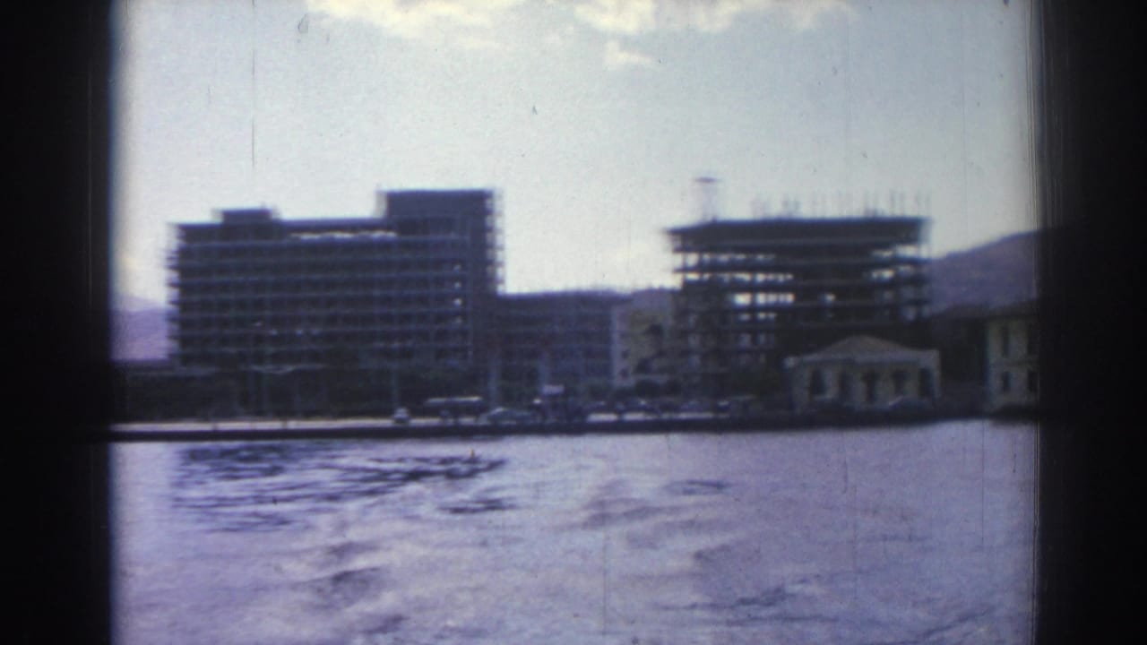 Waterfront buildings and construction seen across rippling water under a cloudy sky.