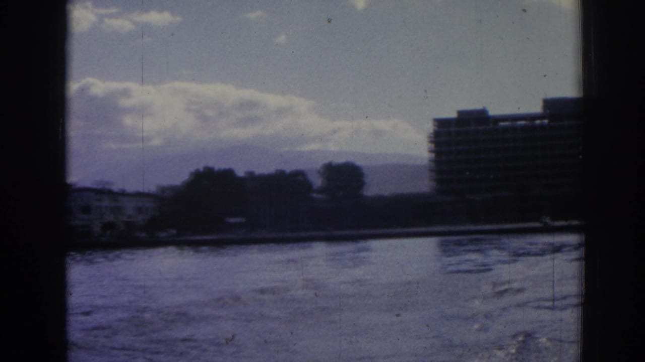 1962 Izmir waterfront pan with water in front, trees along the shore, a cropped high-rise, clouds, and distant hills.