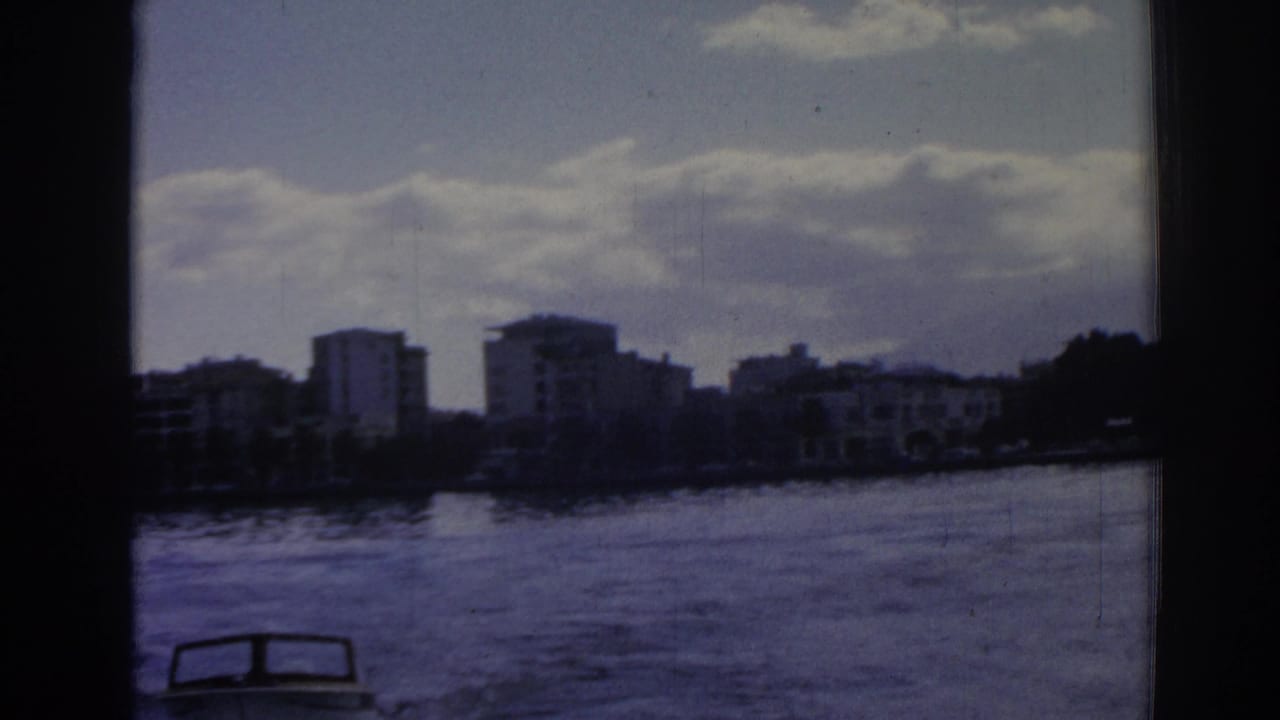 Small motorboat at the lower edge of choppy water with distant shoreline buildings under clouds.