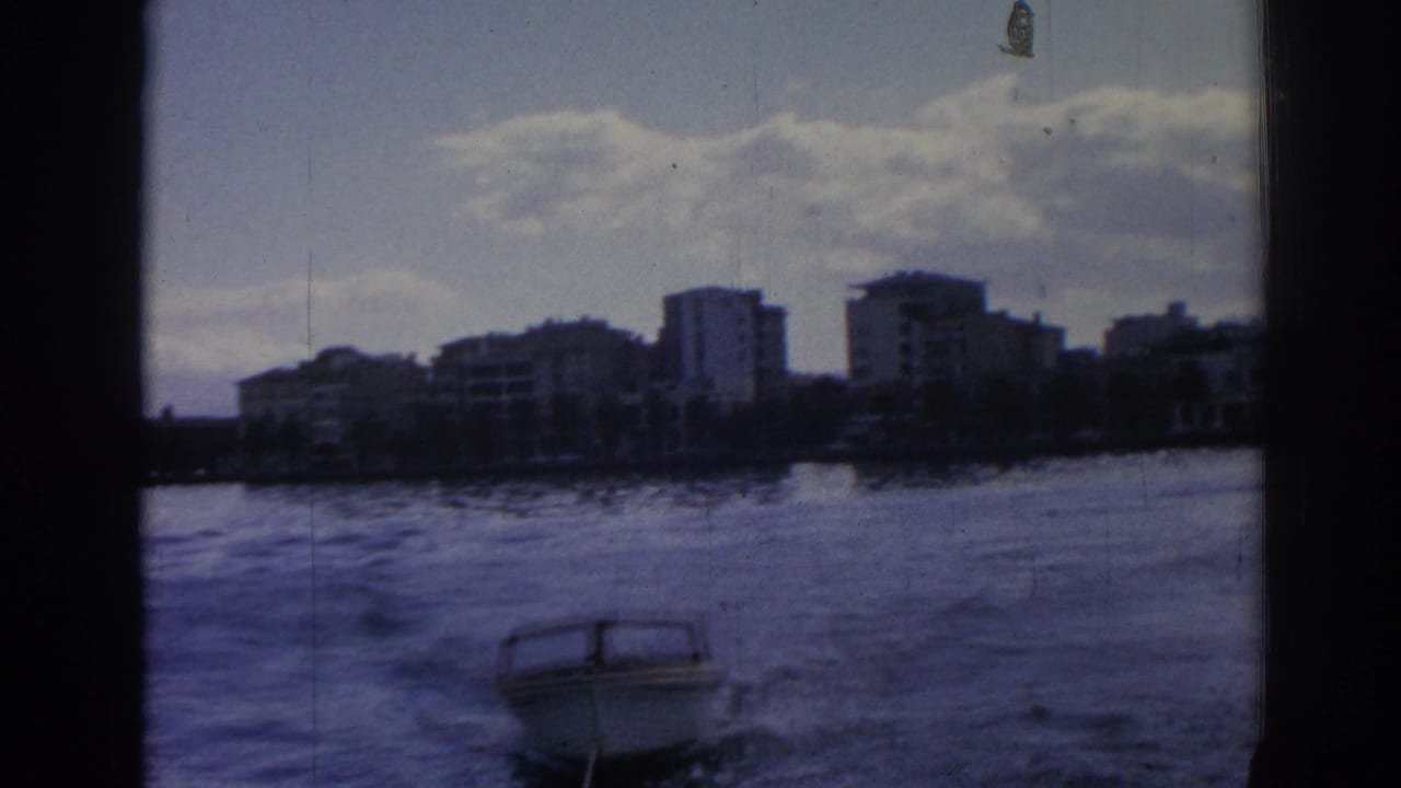 1962 Izmir waterfront view from a moving vessel, with a small motorboat on rippled water and mid-rise buildings on the shore.