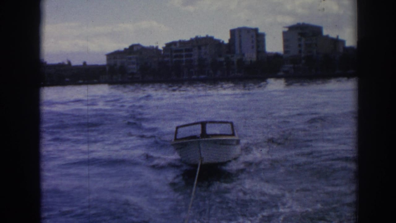 A 1962 8mm frame from Izmir shows a small boat following through wake, with choppy water and an urban shoreline in the background.
