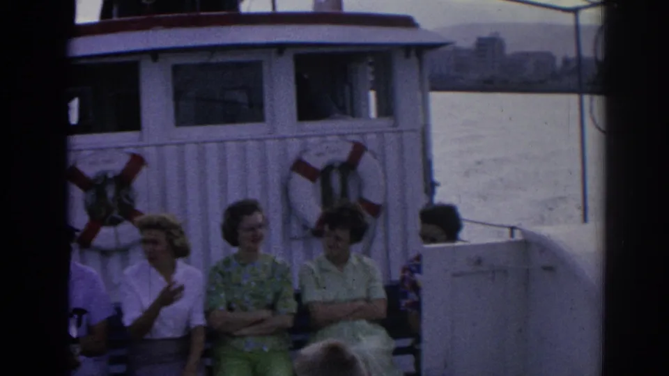 1962 Izmir ferry deck scene with adults seated on benches beside life rings and a white cabin, with water behind them.